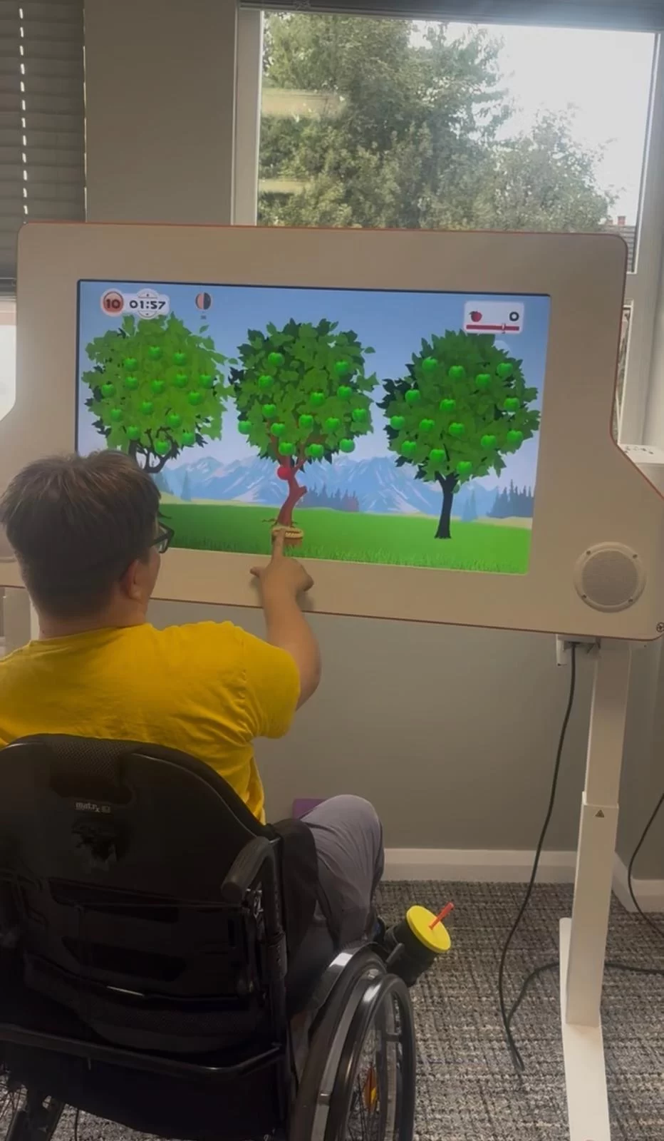 A young person in a wheelchair reaches out to pick an apple from a tree in a touch-screen game