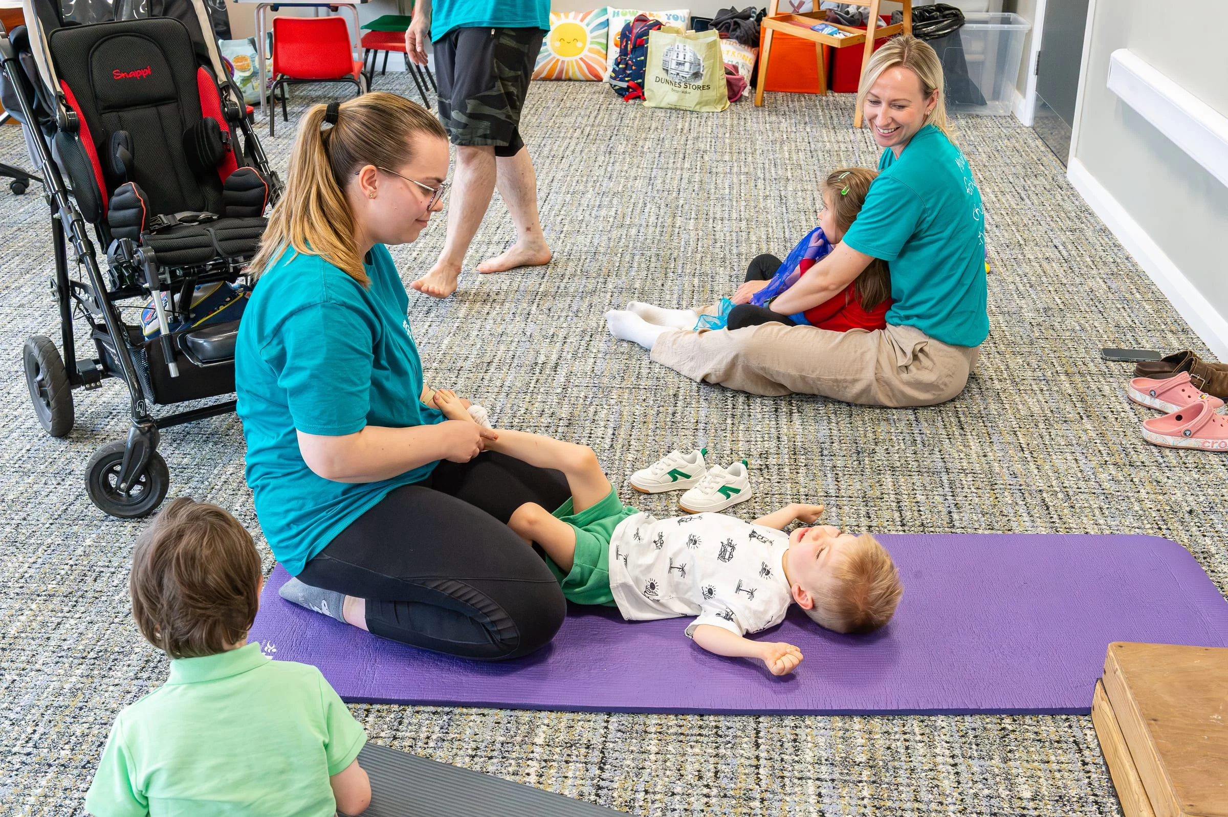 A therapist working with a baby on a padded mat, with a wheelchair and other children nearby