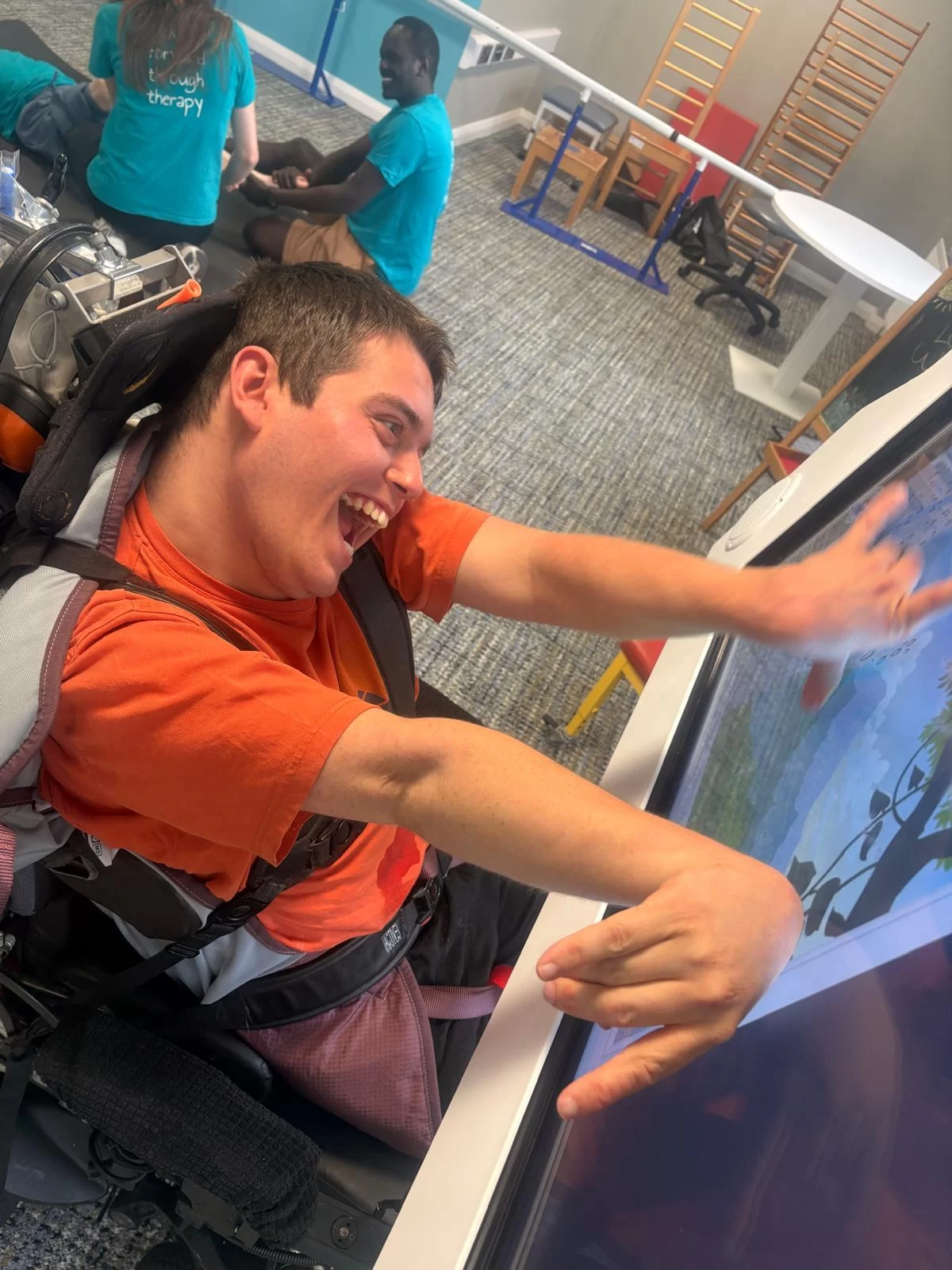 A young person in a wheelchair laughs while stretching both arms across a large touch screen