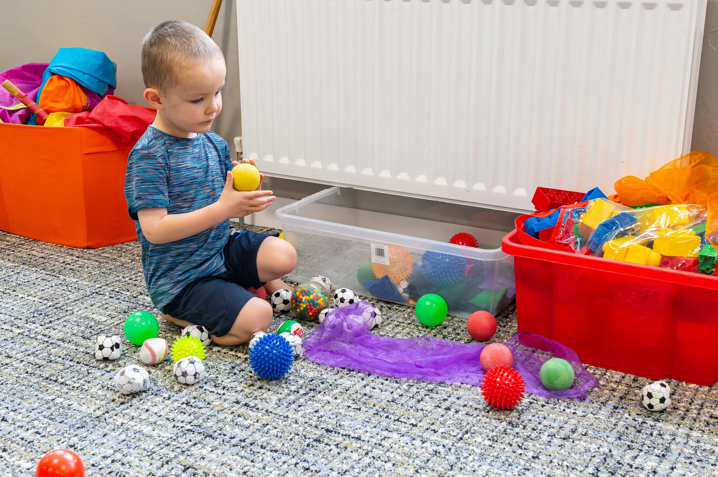 A young child kneeling on the floor, selecting a ball from a scattered set of coloured sensory balls