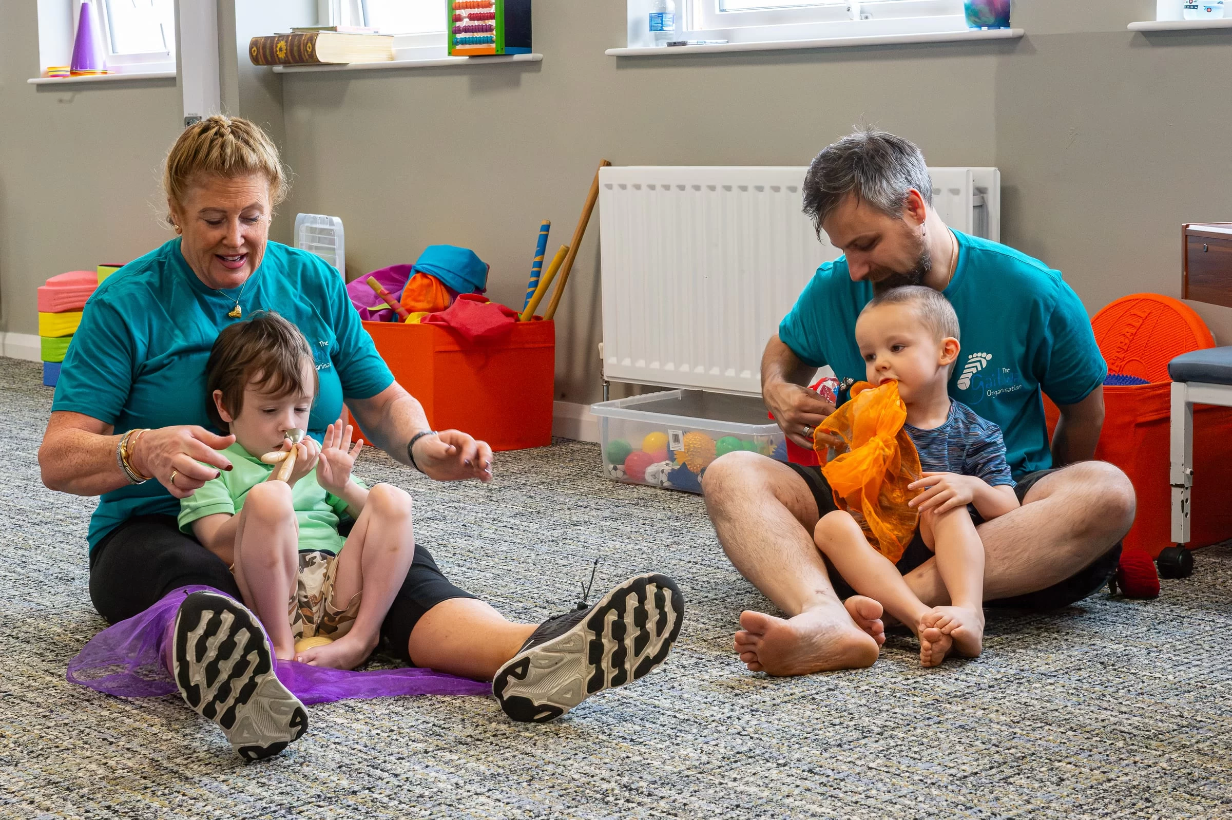 Two therapists working with young children on the floor, using brightly coloured sensory scarves