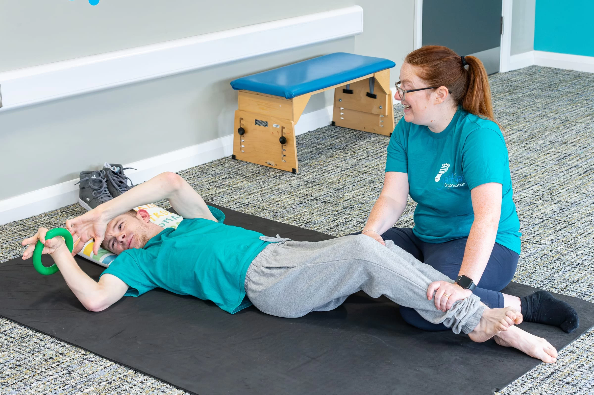 A therapist guiding a young person through a supported leg stretch on a floor mat