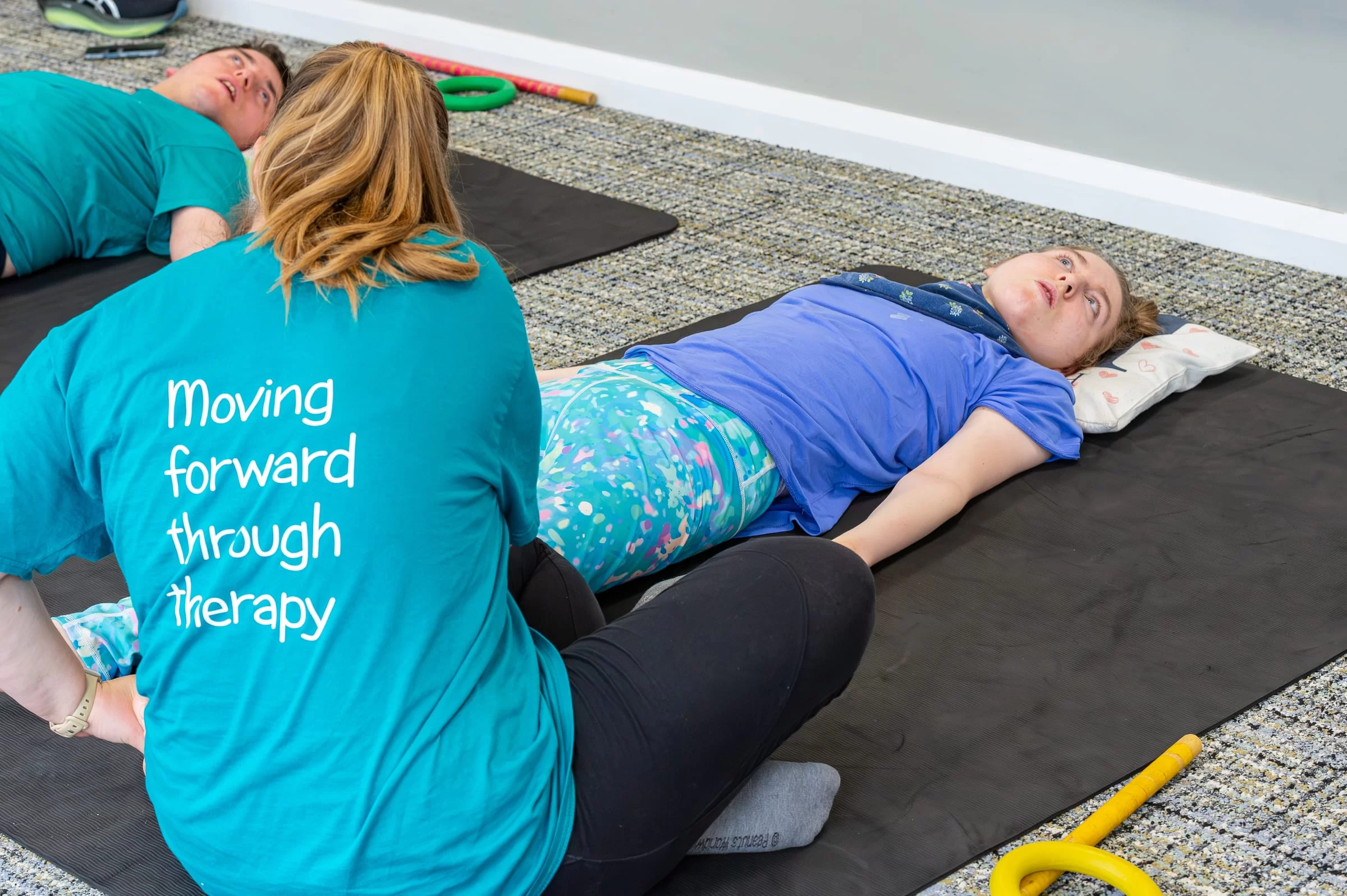 A therapist working at a mat, the Gaitway strapline 'Moving forward through therapy' visible on the back of the shirt