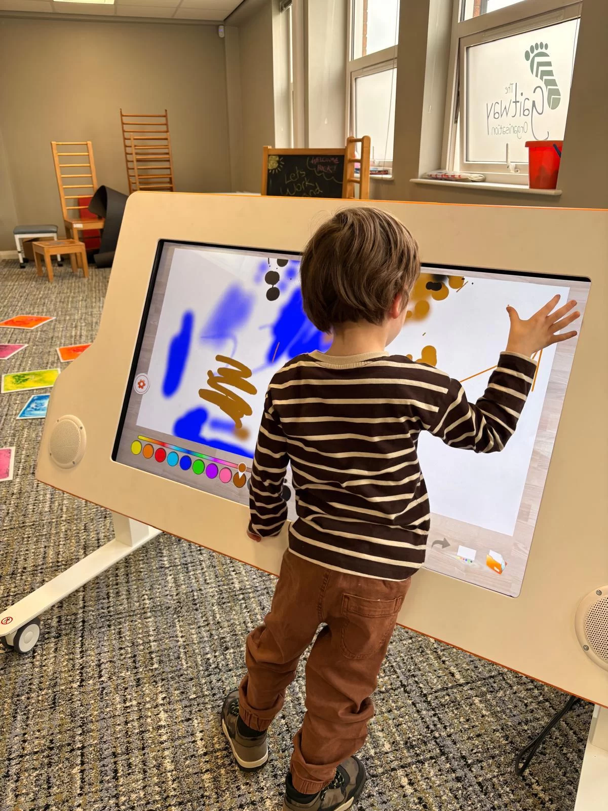 A young boy stands at a large tilted touch screen, painting in bright colours with his hand