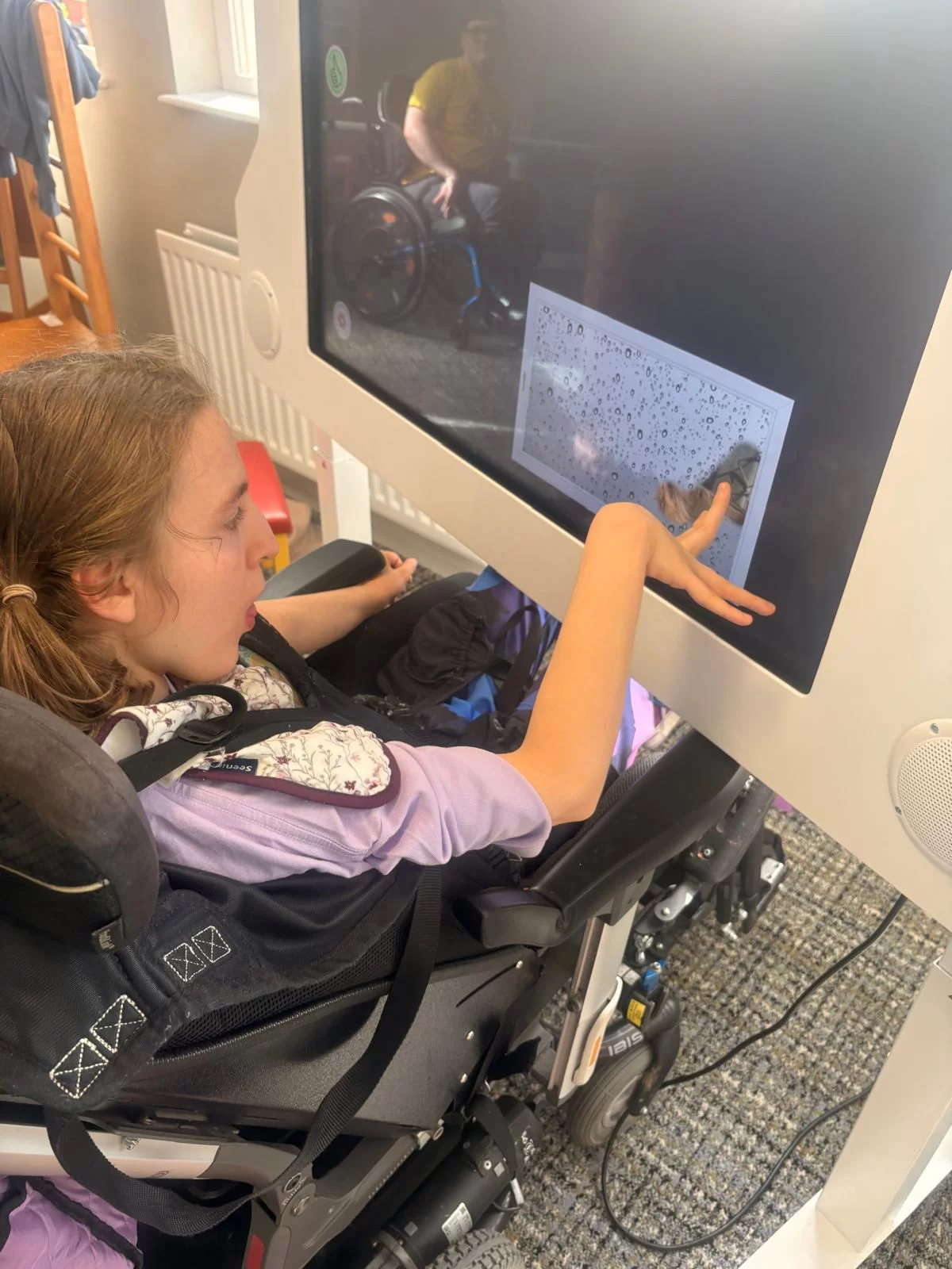 A girl in a wheelchair reaches out to touch a picture of raindrops on a large touch screen