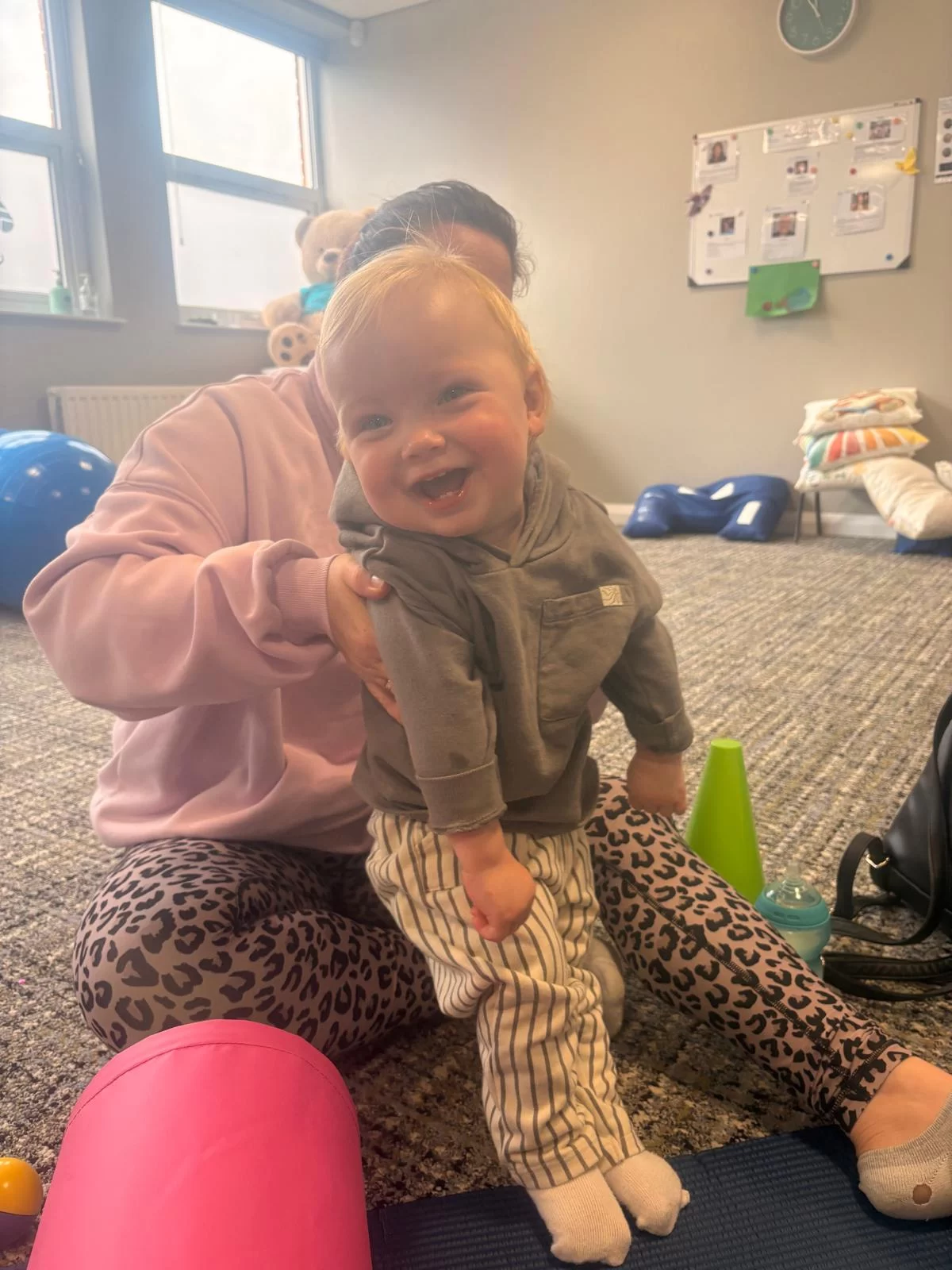 A smiling toddler stands on a mat, held steady at the shoulders by an adult sitting behind