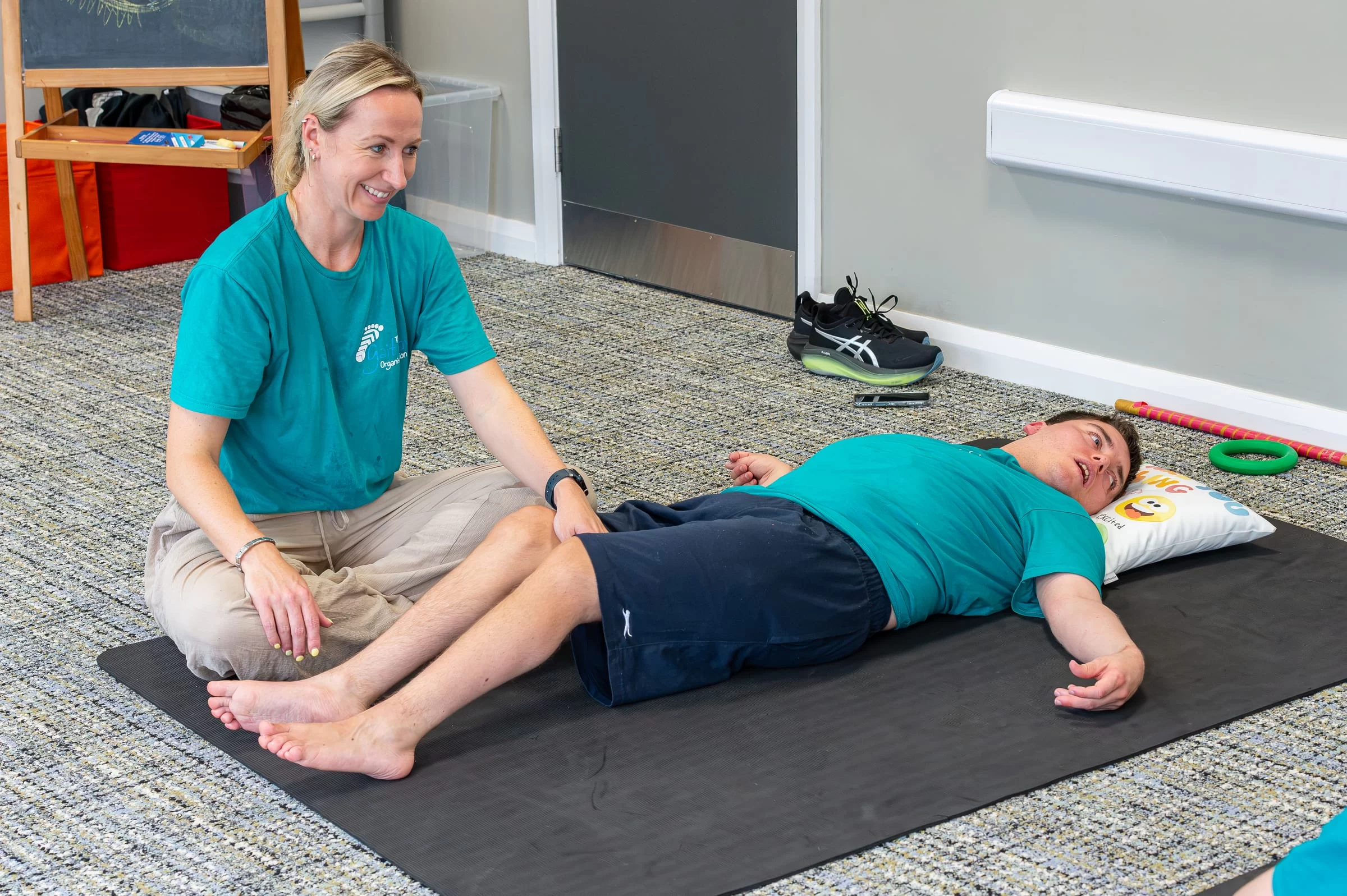 A therapist sitting cross-legged on a mat, smiling while working with a young person