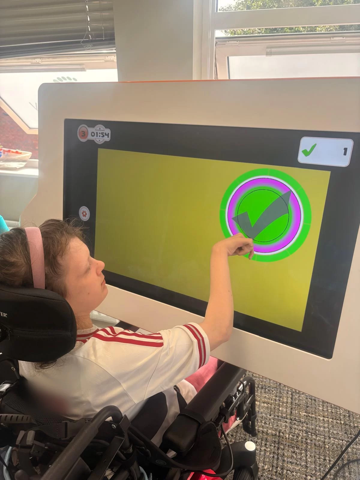 A young person in a wheelchair points at a target on a large touch screen showing a green tick