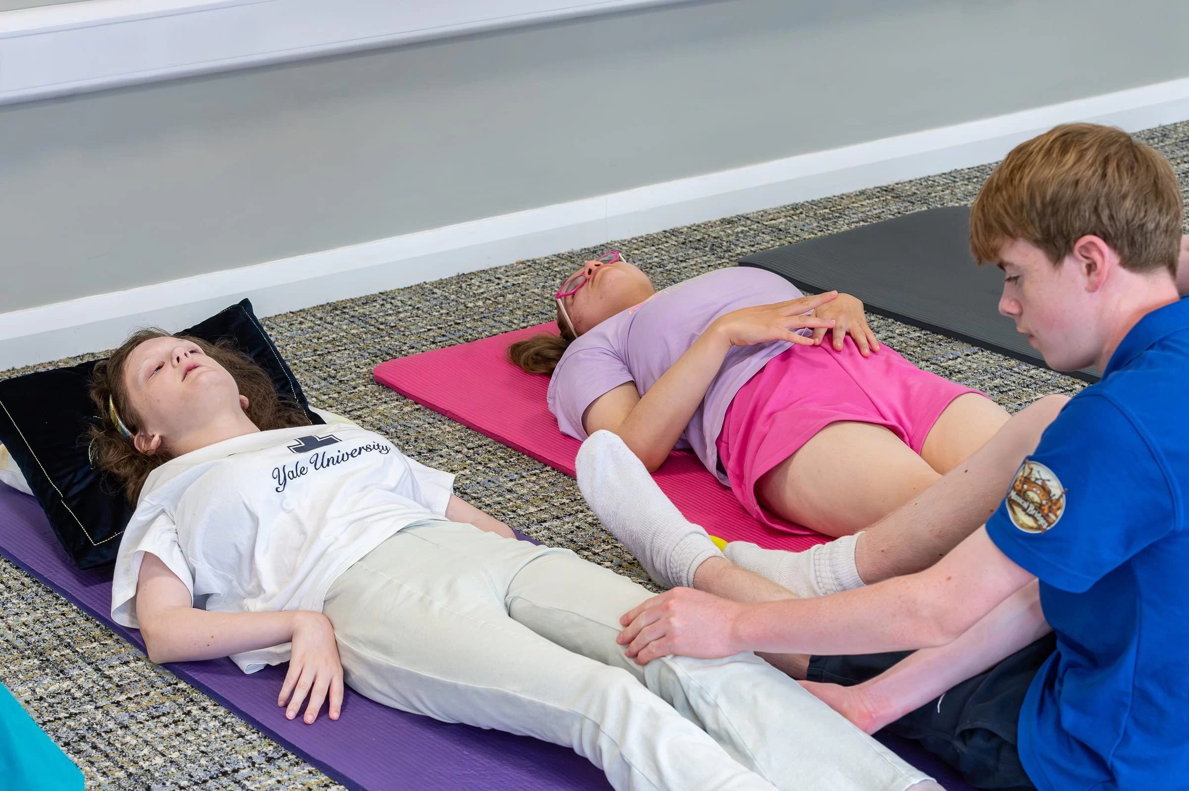 Two young people resting on mats with a helper alongside during a group session