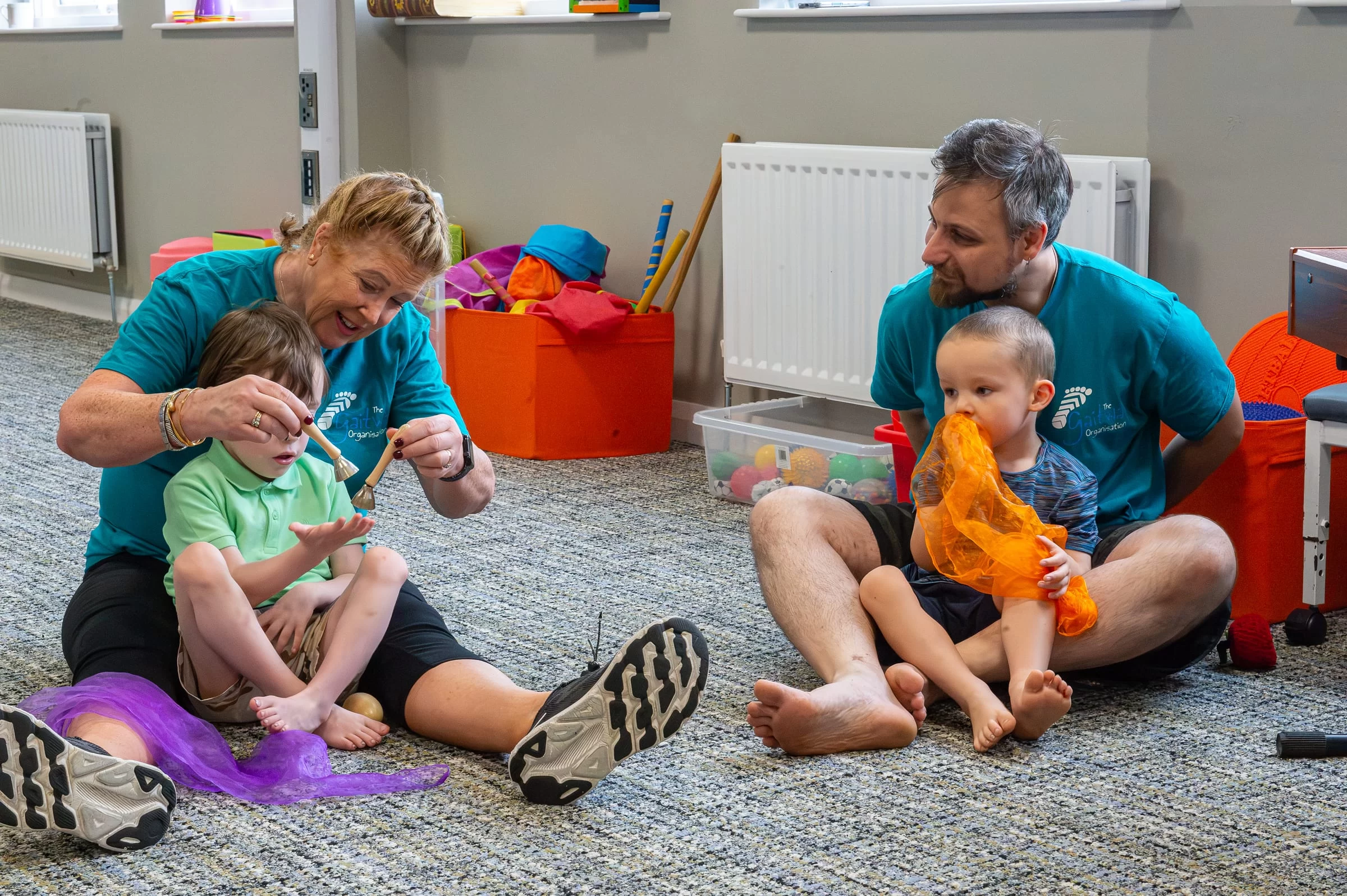 Two therapists seated on the floor, each supporting a young child during a play-based session