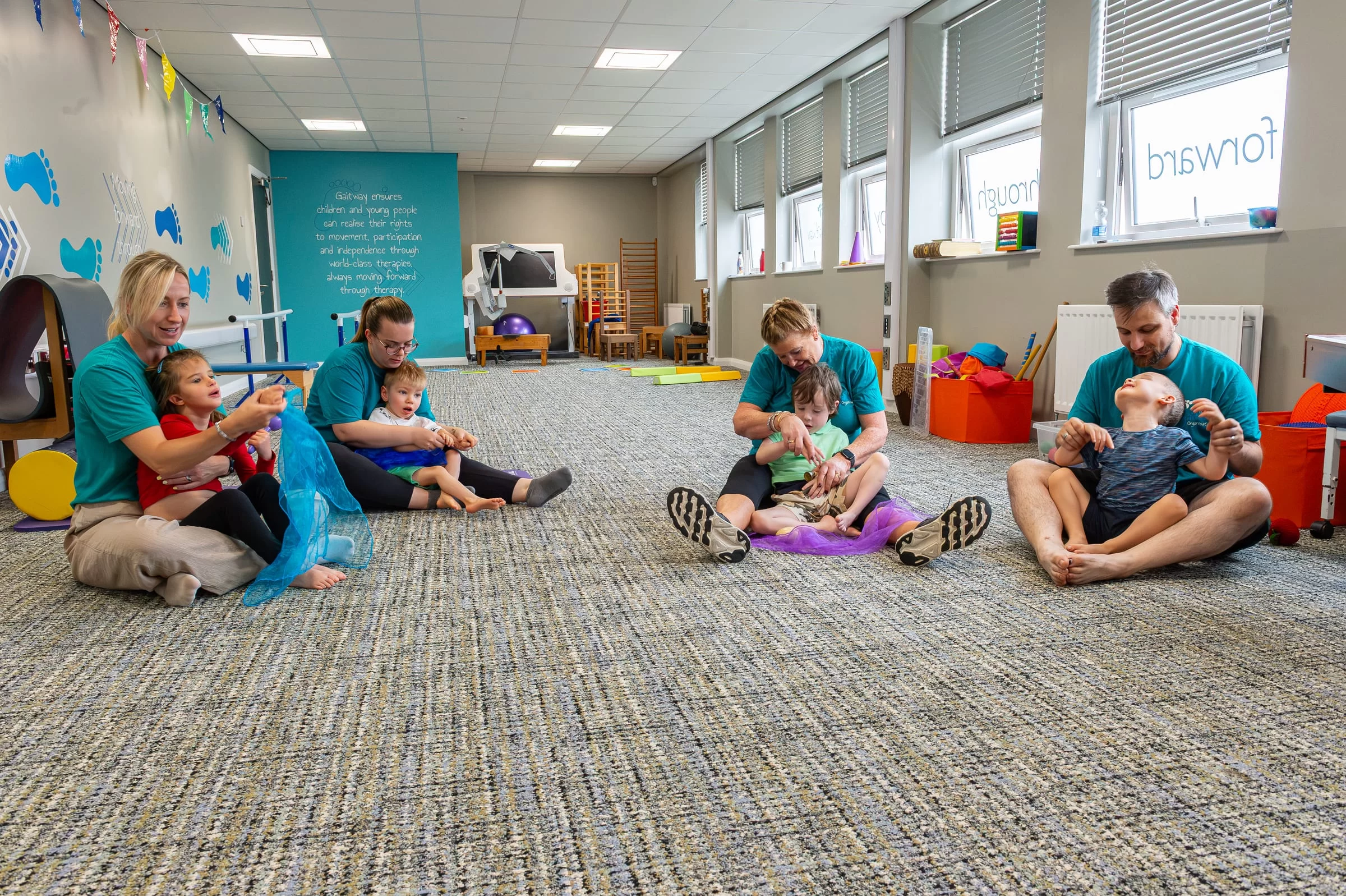 A wide view of the Gaitway therapy room, with several therapists and children working together across the floor