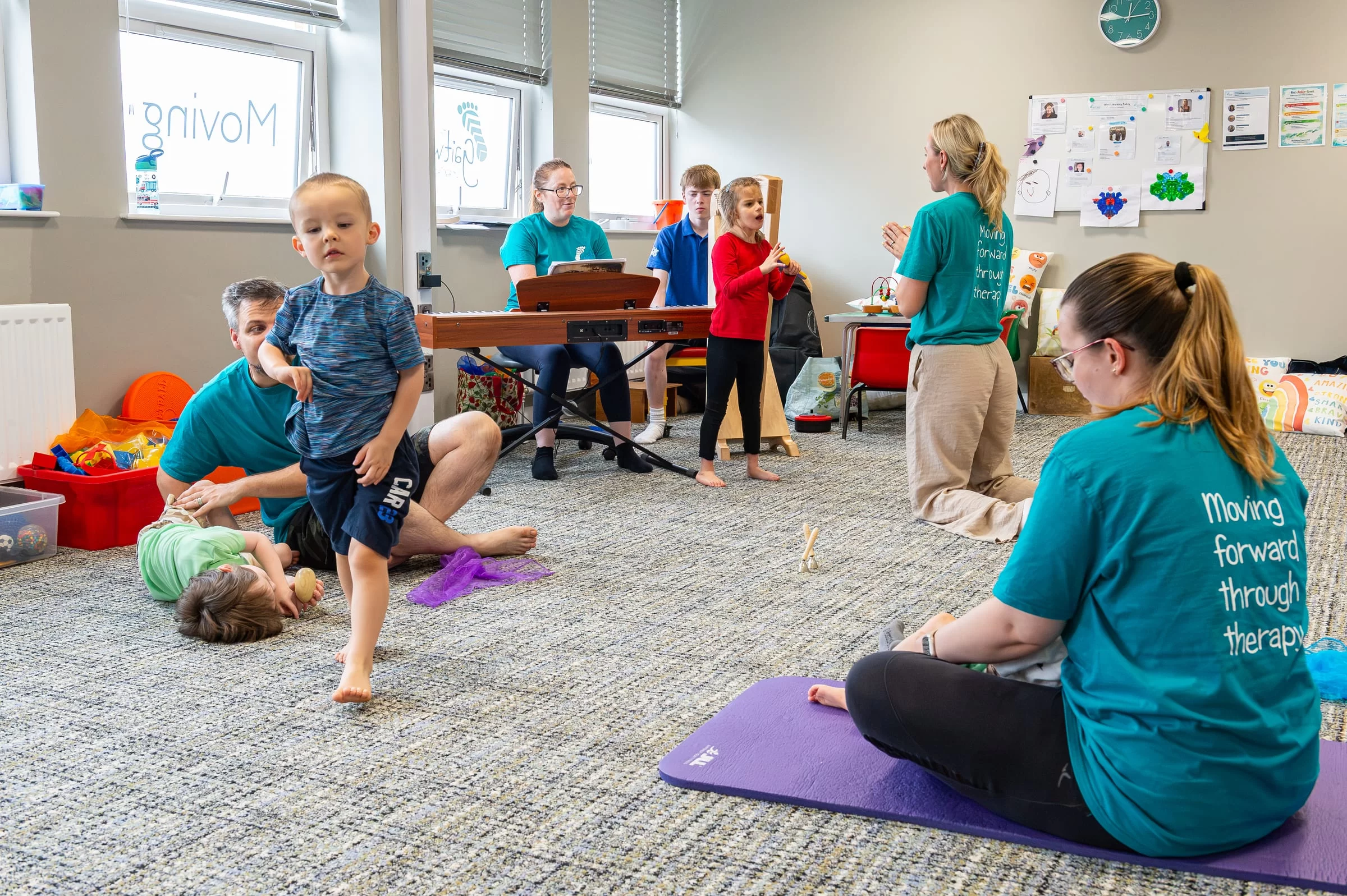 A child taking supported steps across the therapy room while live music is played, with therapists and other children nearby