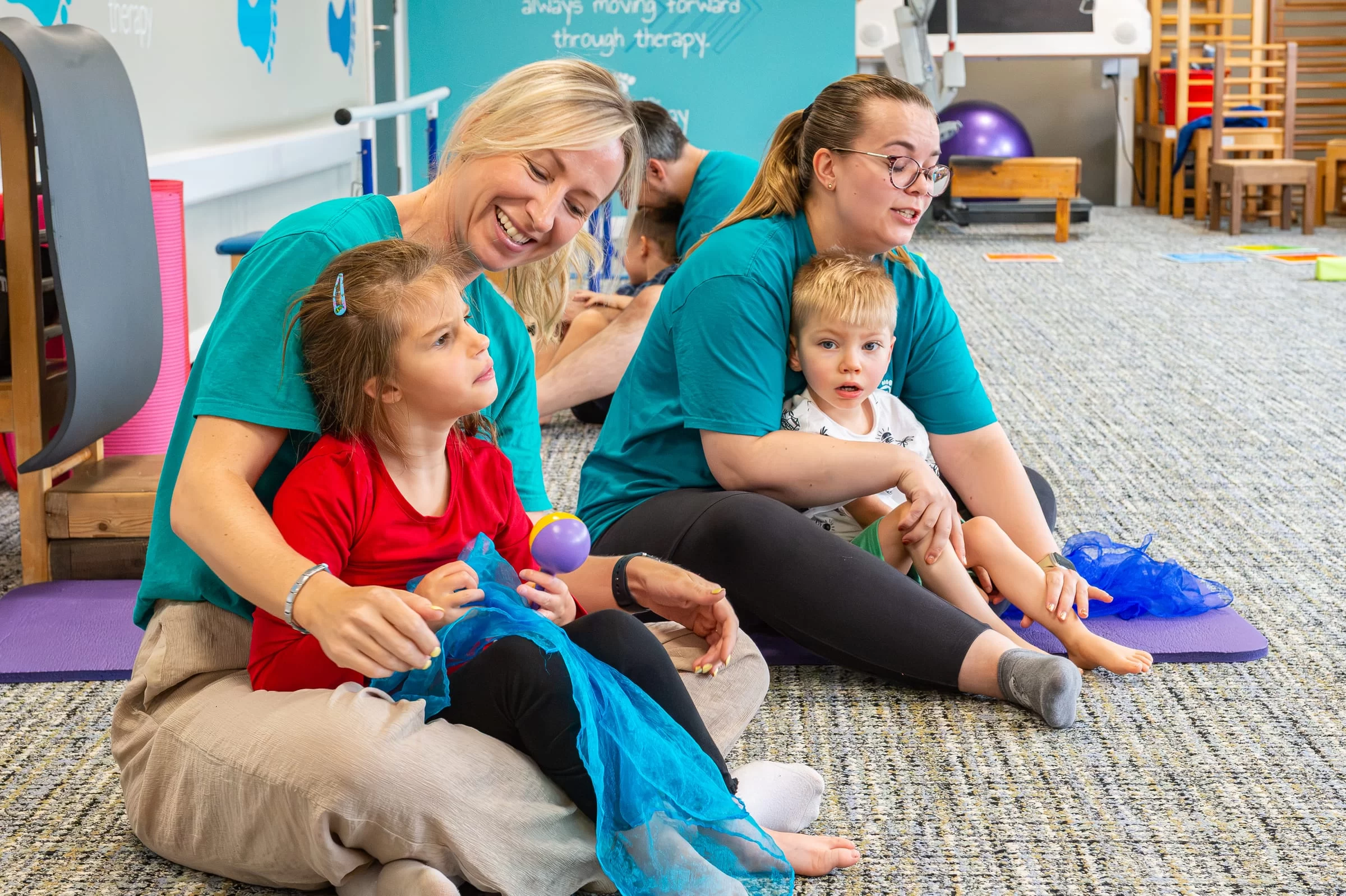 Two therapists sitting on the floor with two young children, all smiling, during a sensory play session