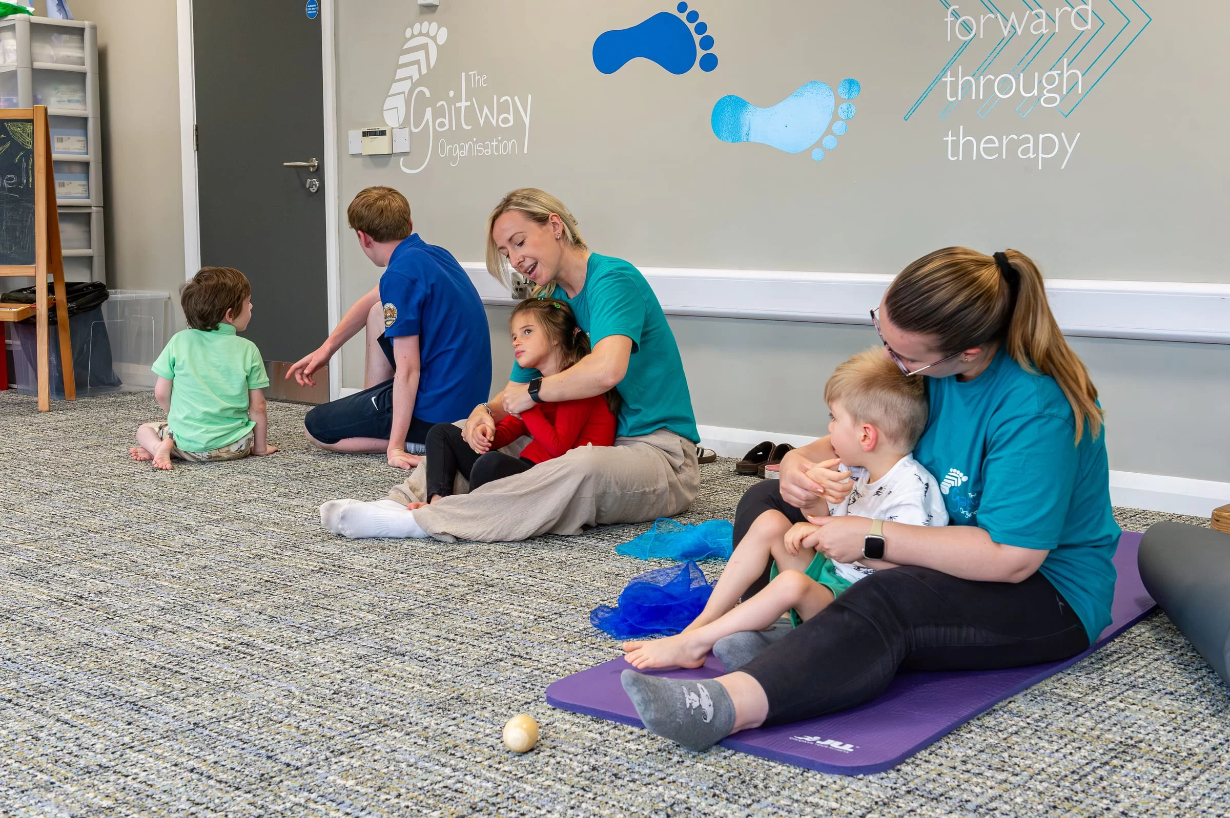 A therapist sitting on a mat with a young child during a one-to-one session, the therapy room visible behind