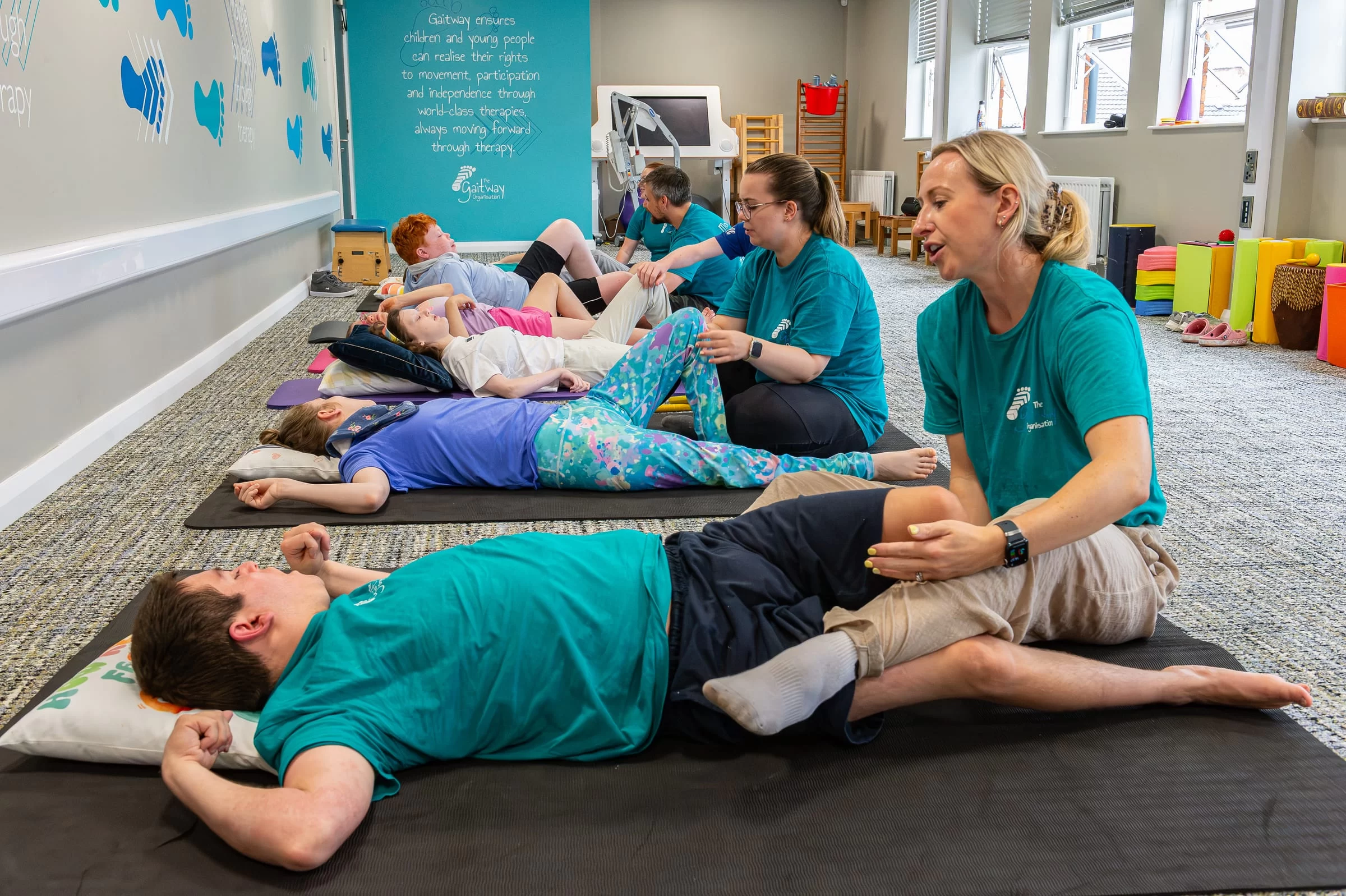 A wide view of a group session, several therapists working one-to-one with children on floor mats beneath the Gaitway strapline wall