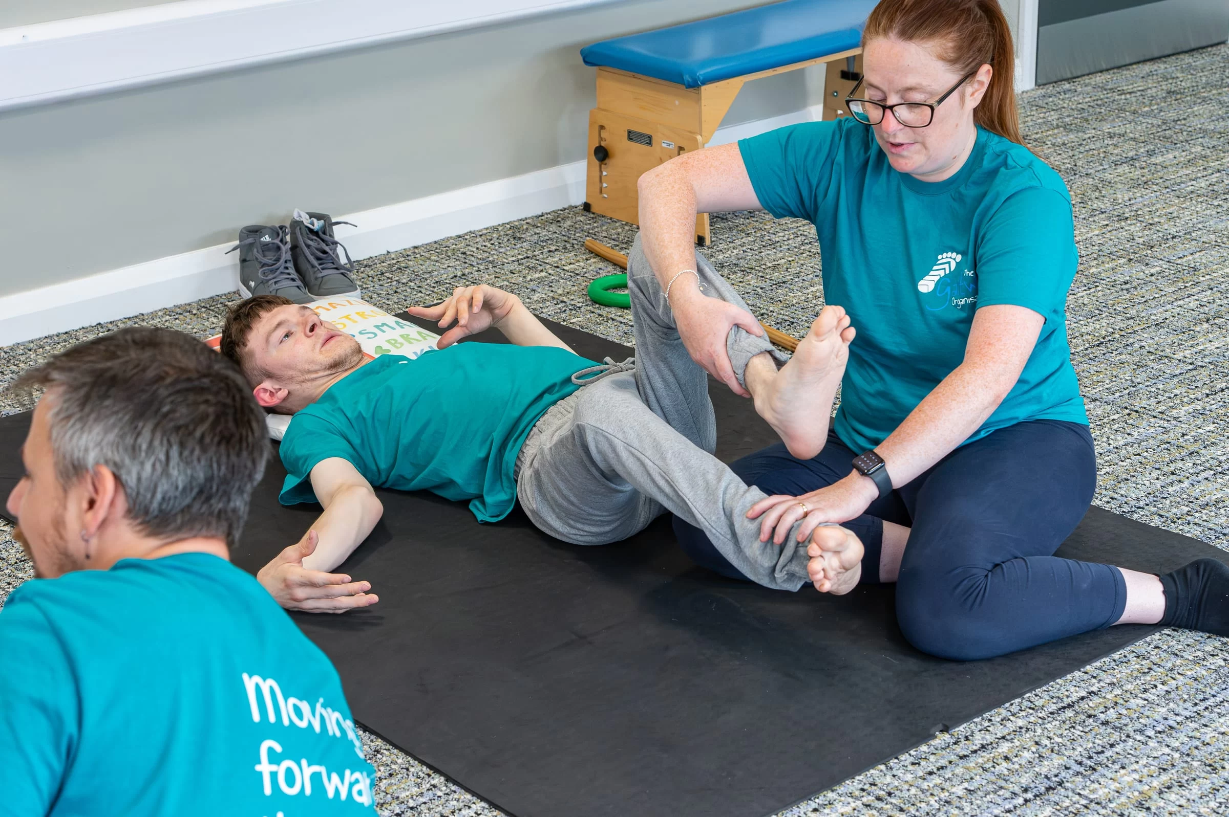 A therapist guiding a young person through a supported leg stretch on a floor mat