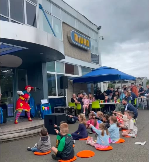 A family fun day outside the Mivan building, with children seated on the ground watching an entertainer