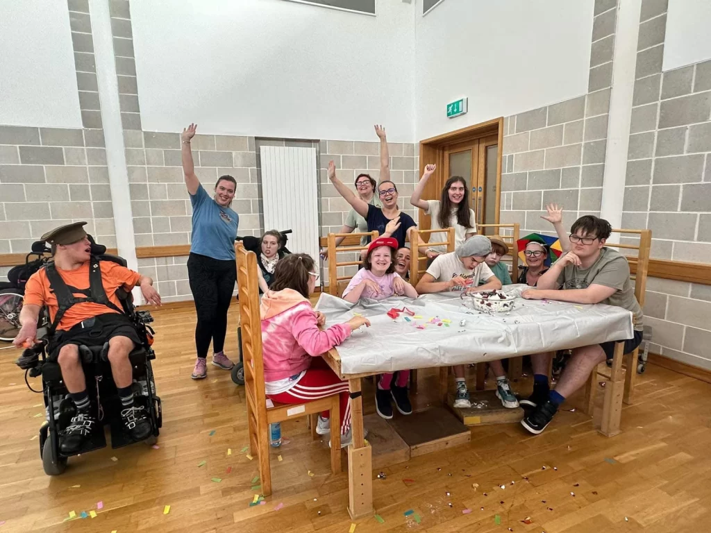 Children and staff around a craft table in the therapy room, several waving at the camera
