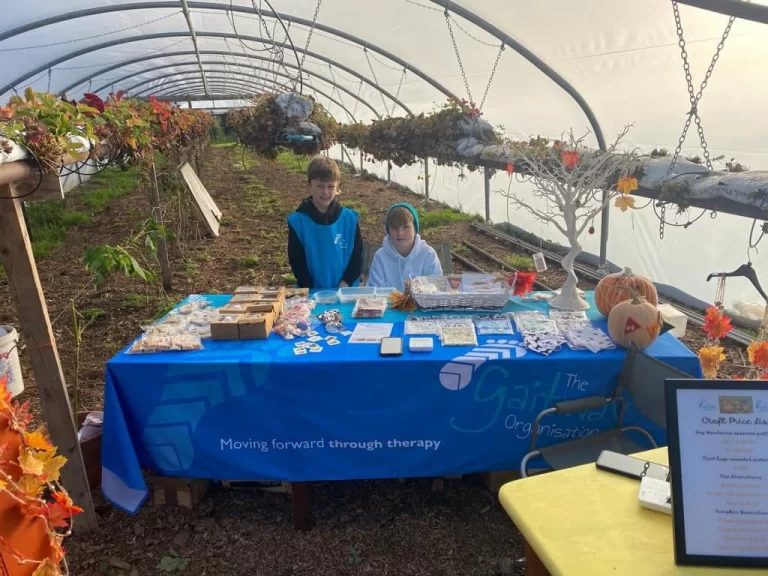 Two children behind the Gaitway craft stall in a polytunnel at the pumpkin patch, its blue table cover reading "Moving forward through therapy"