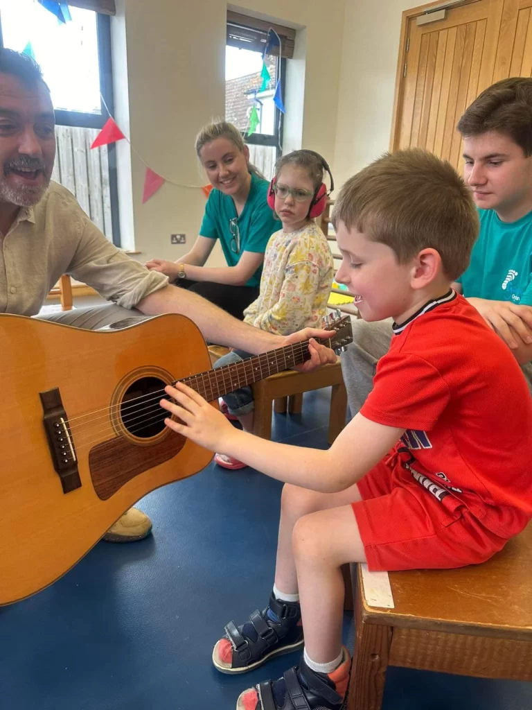 A child playing an acoustic guitar with support from an adult during a music therapy session